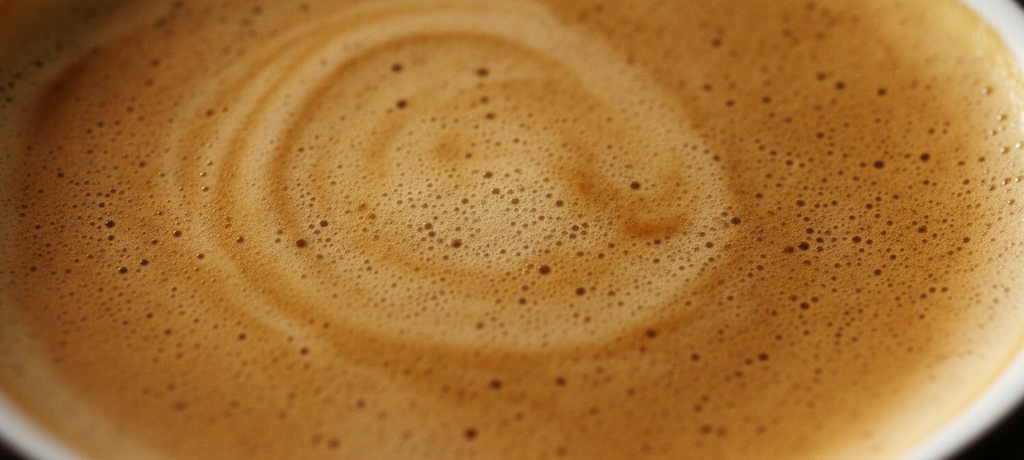 A close-up artistic shot of rich velvety coffee crema swirling in a cup, capturing dense golden-brown foam texture with subtle ripples, warm soft lighting highlighting the creamy layers, evoking a sense of indulgence and craftsmanship